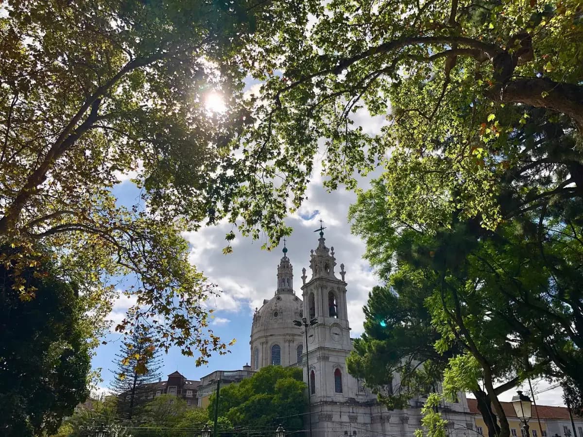 A white stone church with a large dome and an ornate bell tower rises behind trees, seen from a park under a canopy of leaves against a cloudy blue sky.
