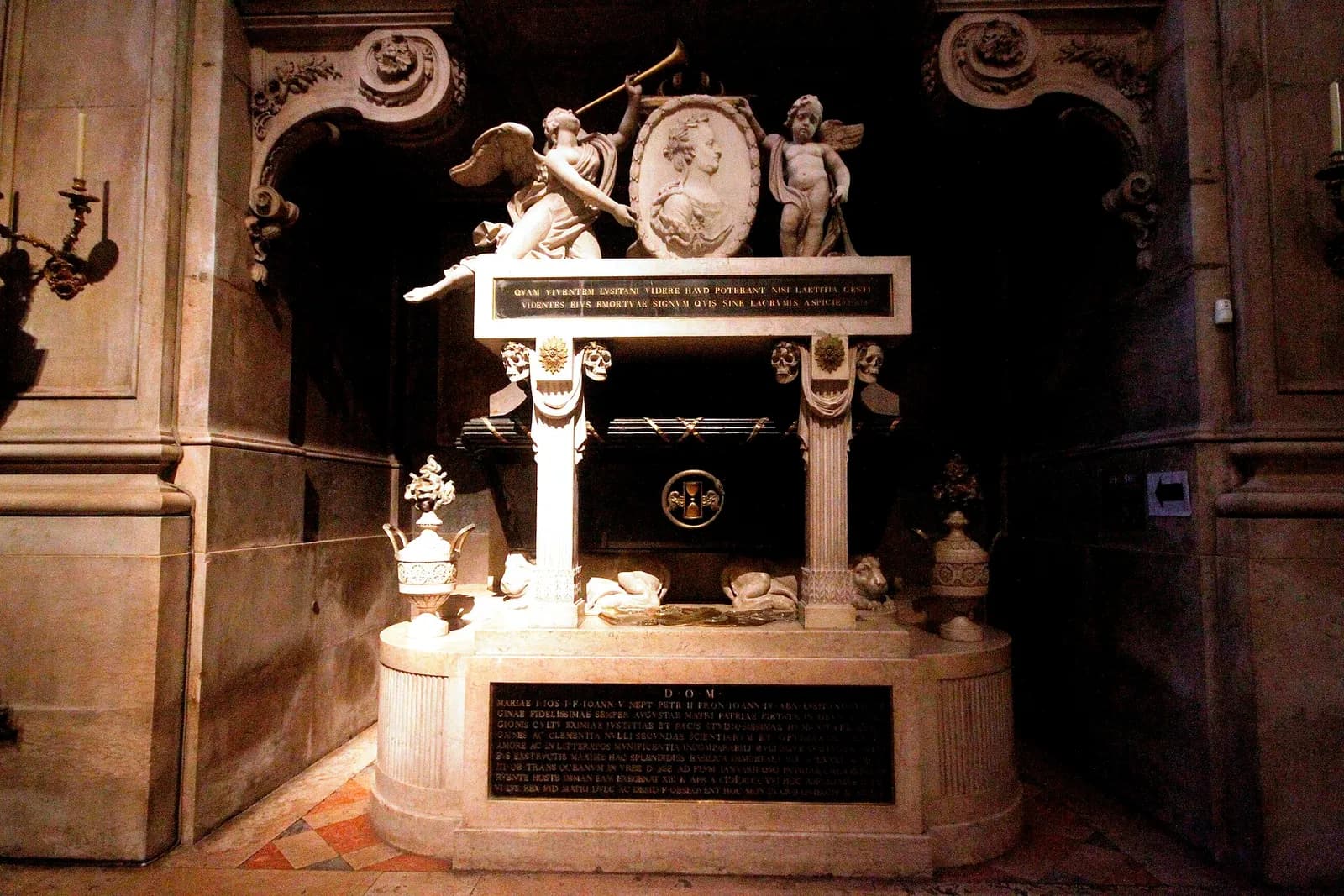 A marble tomb in a dim church transept: a dark sarcophagus between two columns, topped by a medallion portrait of a woman in profile flanked by a trumpeting angel and a cherub, with Latin inscriptions in gold on black panels.