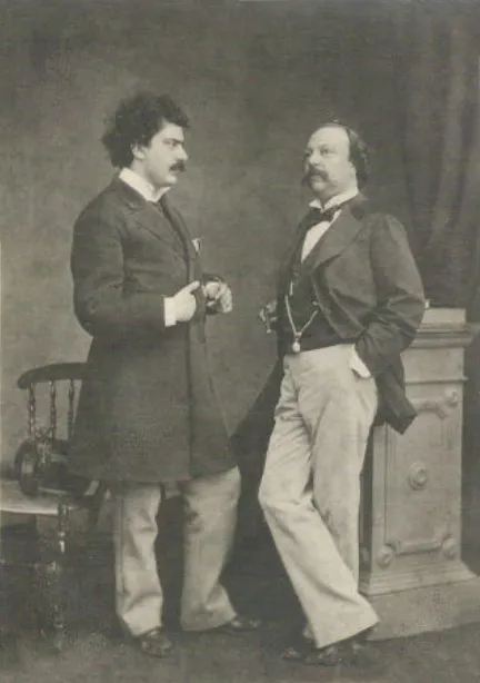 A studio photograph of two men in frock coats and pale trousers. On the left a younger man with thick curly hair and a moustache holds his lapel; on the right an older, heavier man with side whiskers leans on a pedestal.