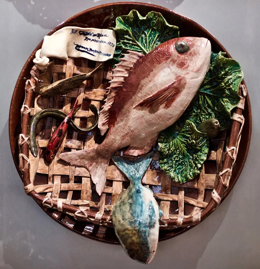 A round brown glazed wall plate modelled in relief as a woven wicker tray. A large pink fish lies across it on green cabbage leaves, with smaller fish and an eel beside it, and a white ribbon at the top bears a painted inscription.