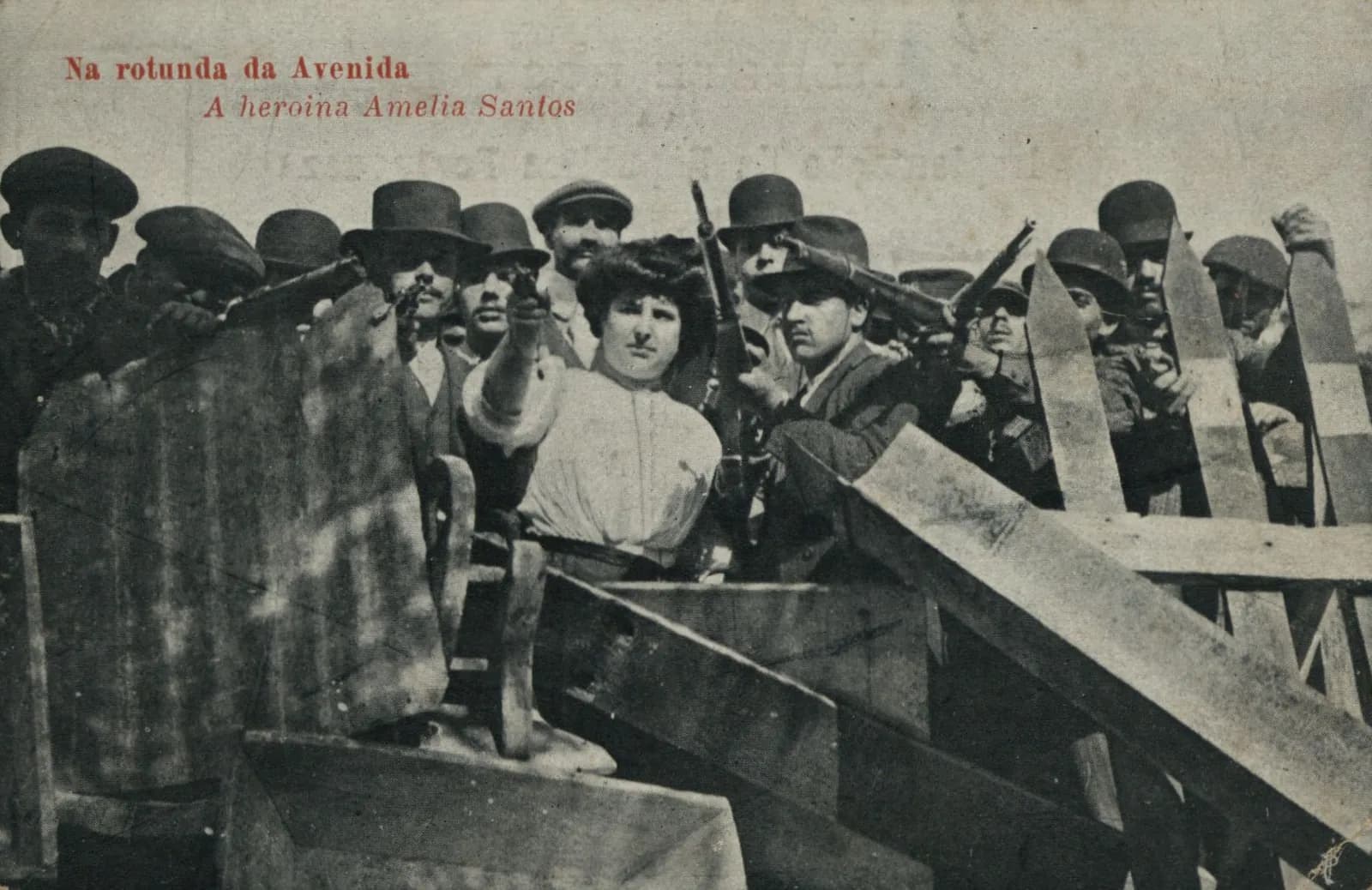 A postcard photograph of a barricade of planks. A woman in a pale blouse stands among men in caps and bowler hats, pointing a pistol forward, while the men around her hold rifles. Red lettering at the top reads Na rotunda da Avenida, A heroina Amelia Santos.