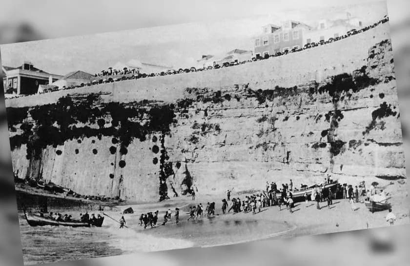 A beach under a high rock cliff. A group of people walks to the water's edge towards a rowing boat, other boats lie on the sand, and spectators line the top of the cliff in front of a row of houses.