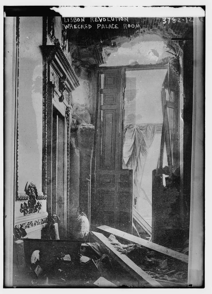 A palace room with a shattered wall and doorway, loose planks and debris on the floor, a torn curtain hanging and an ornate carved frame on the left. Handwritten text at the top reads Lisbon Revolution wrecked palace room.