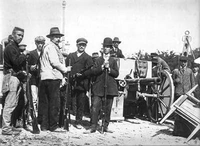 A group of men, some in caps and soldiers' uniforms, others in civilian suits and bowler hats, stand holding rifles beside a field gun on open ground.