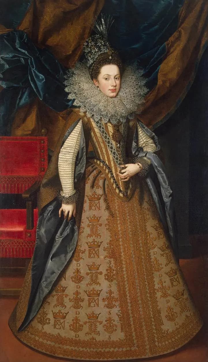 Full-length portrait of a young woman with a pale face and a towering lace ruff, wearing a jewelled headdress and a wide gold-embroidered gown patterned with small crowns and monograms, standing beside a red velvet chair.