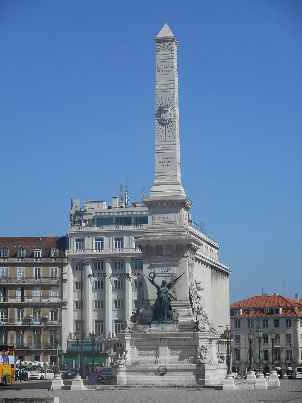 A tall white stone obelisk carved with names and dates rises from a stepped base decorated with a dark bronze winged figure holding a wreath, set in a paved square in front of tall city buildings under a deep blue sky.