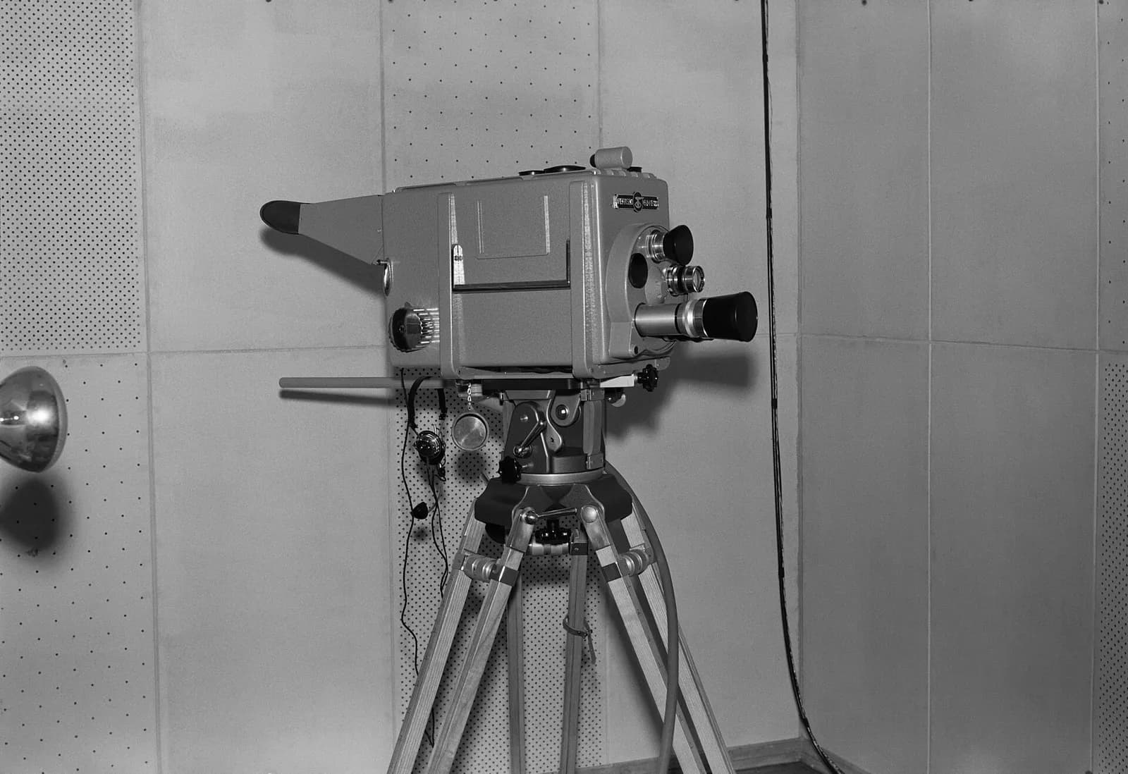 Black-and-white photograph of a boxy television studio camera on a wooden tripod, with a turret of lenses on the front and a long viewing hood at the back, standing in a room with perforated acoustic panels.