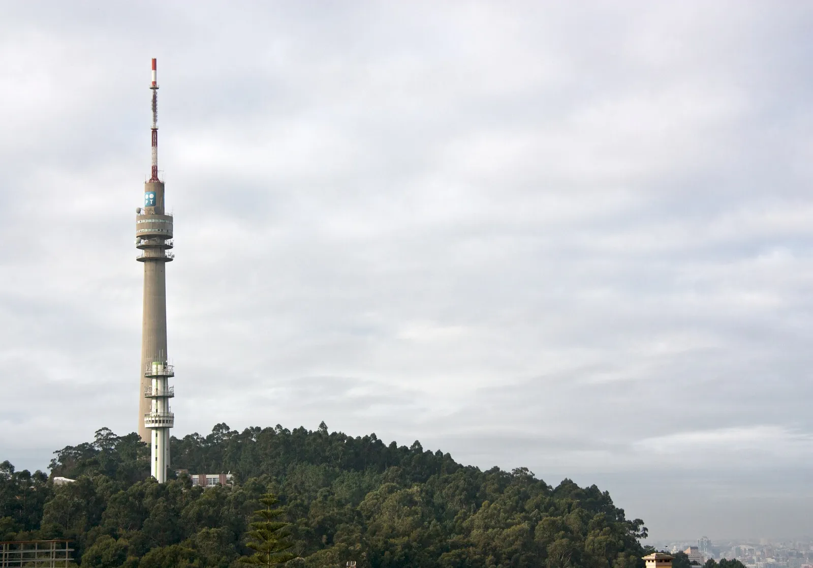 A tall concrete telecommunications tower with a red-and-white mast on top rises from a wooded hill under a grey sky, with a city in the haze at bottom right.
