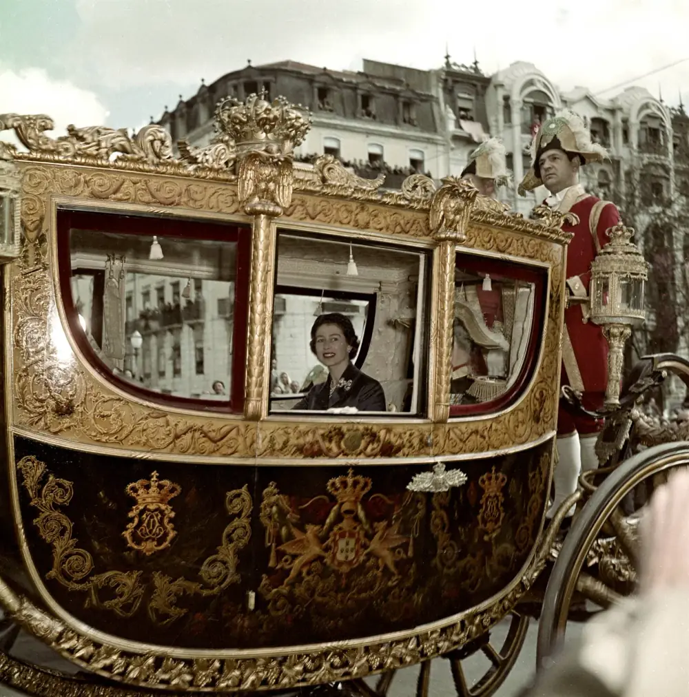 Colour photograph of a young woman in a dark coat and pearls smiling through the window of an ornate gilded coach painted with the Portuguese royal arms, with a footman in a red coat and plumed hat standing at the back and crowded balconies behind.