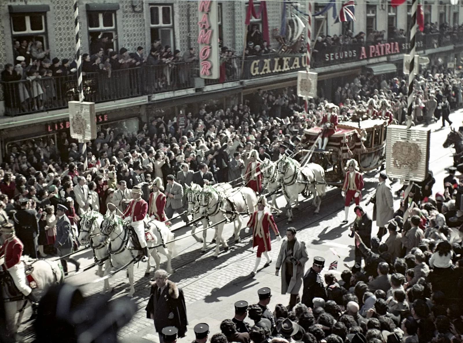 Colour photograph from above of a gilded state coach drawn by white horses, with riders and footmen in red coats, passing along a Lisbon street packed with people. Spectators crowd the pavements and the balconies, where British flags hang beside shop signs.