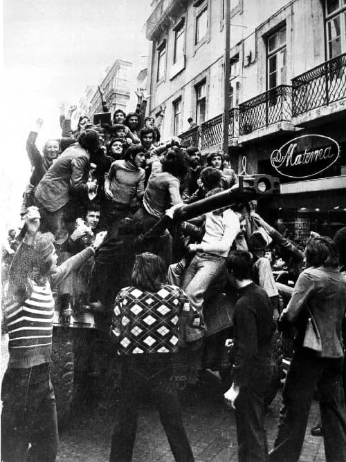 Black-and-white photograph of a crowd of young men and women climbing onto an armoured car in a Lisbon street, raising their arms, in front of a building with wrought-iron balconies and a shop sign reading Materna.
