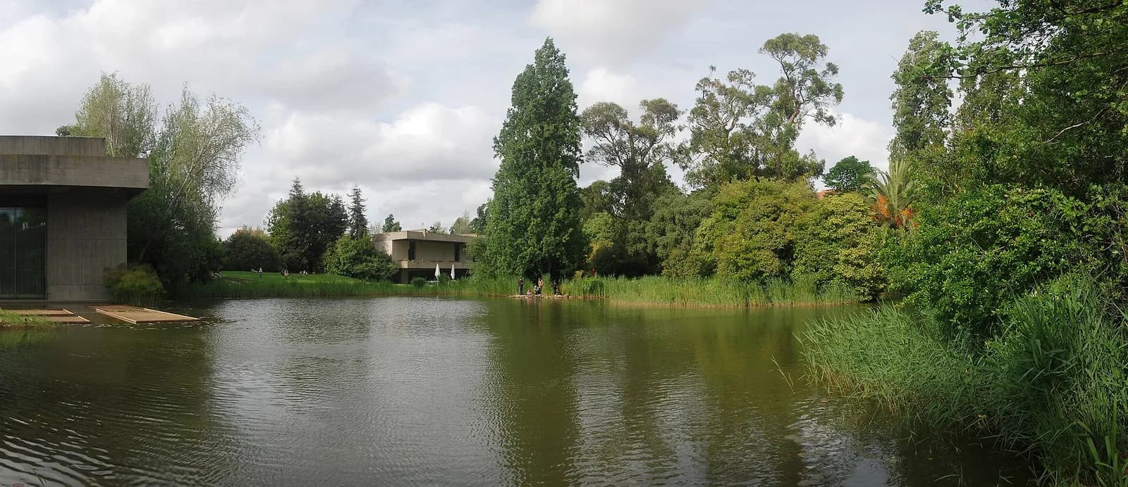 A still lake in a green garden, edged with reeds and tall trees, with a low modern concrete building on the left bank and another among the trees in the distance.
