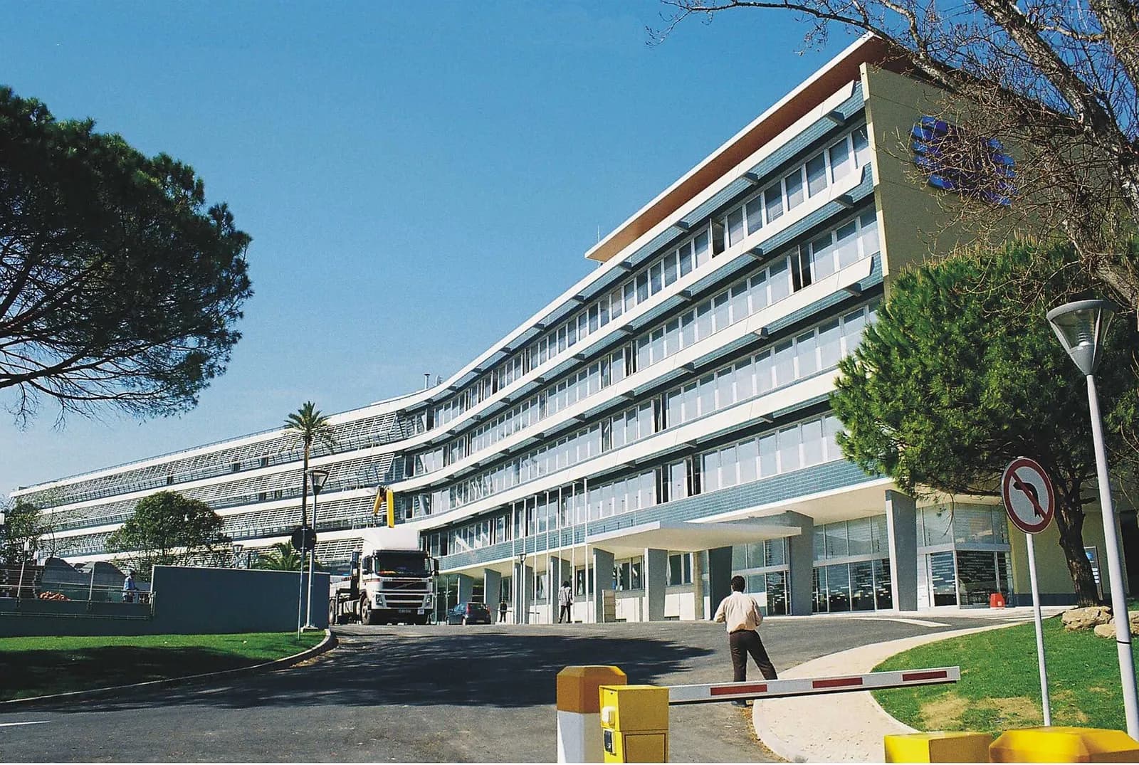 A long, curving modern office block of five storeys with pale walls, rows of windows and a blue logo on its end wall, under a clear blue sky, with pine trees, a lorry and a barrier on the entrance road.