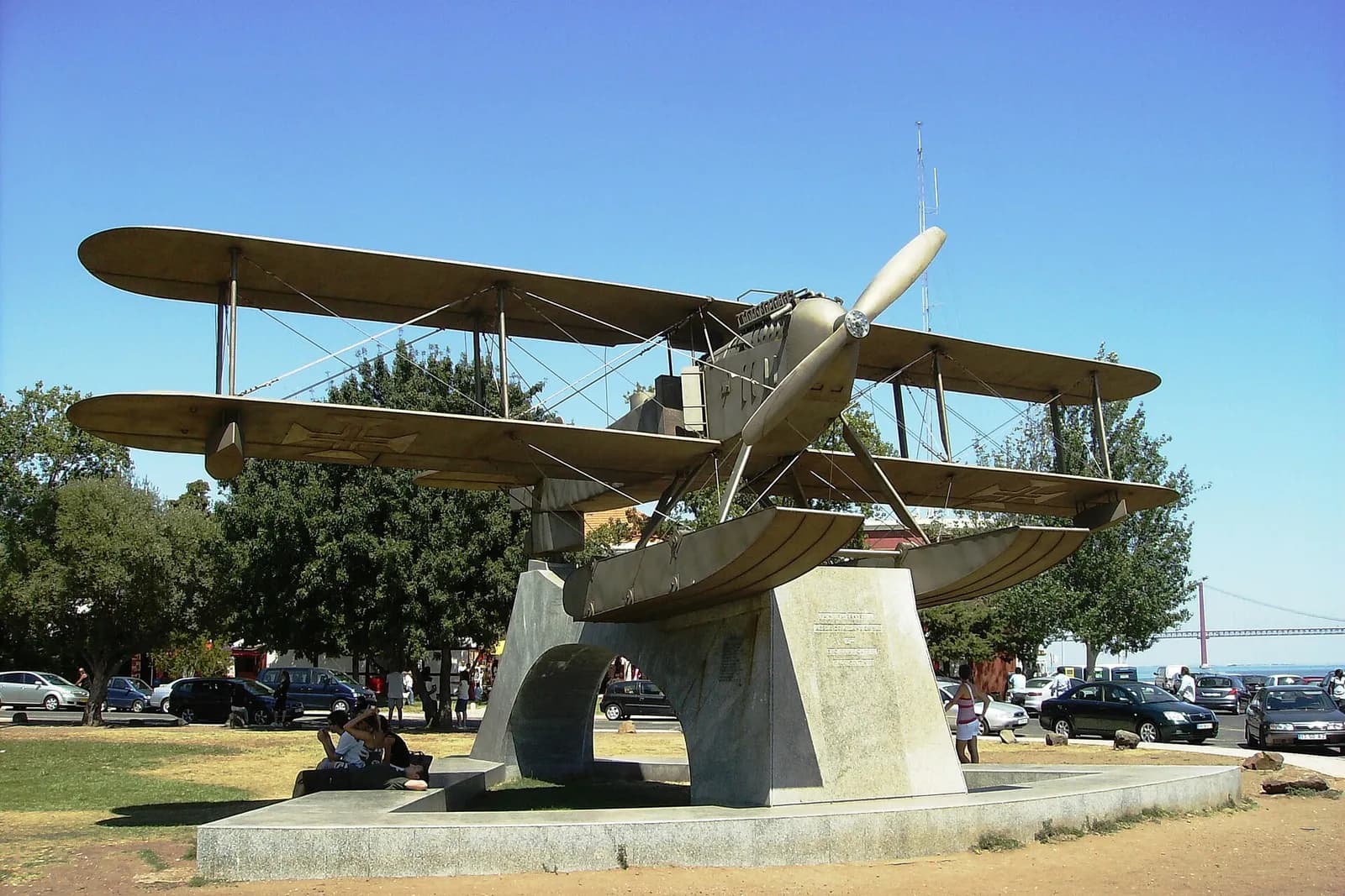 A full-size bronze-coloured metal biplane on floats is mounted on a stone arch in a park under a blue sky, with parked cars behind and the red 25 de Abril suspension bridge in the distance on the right.