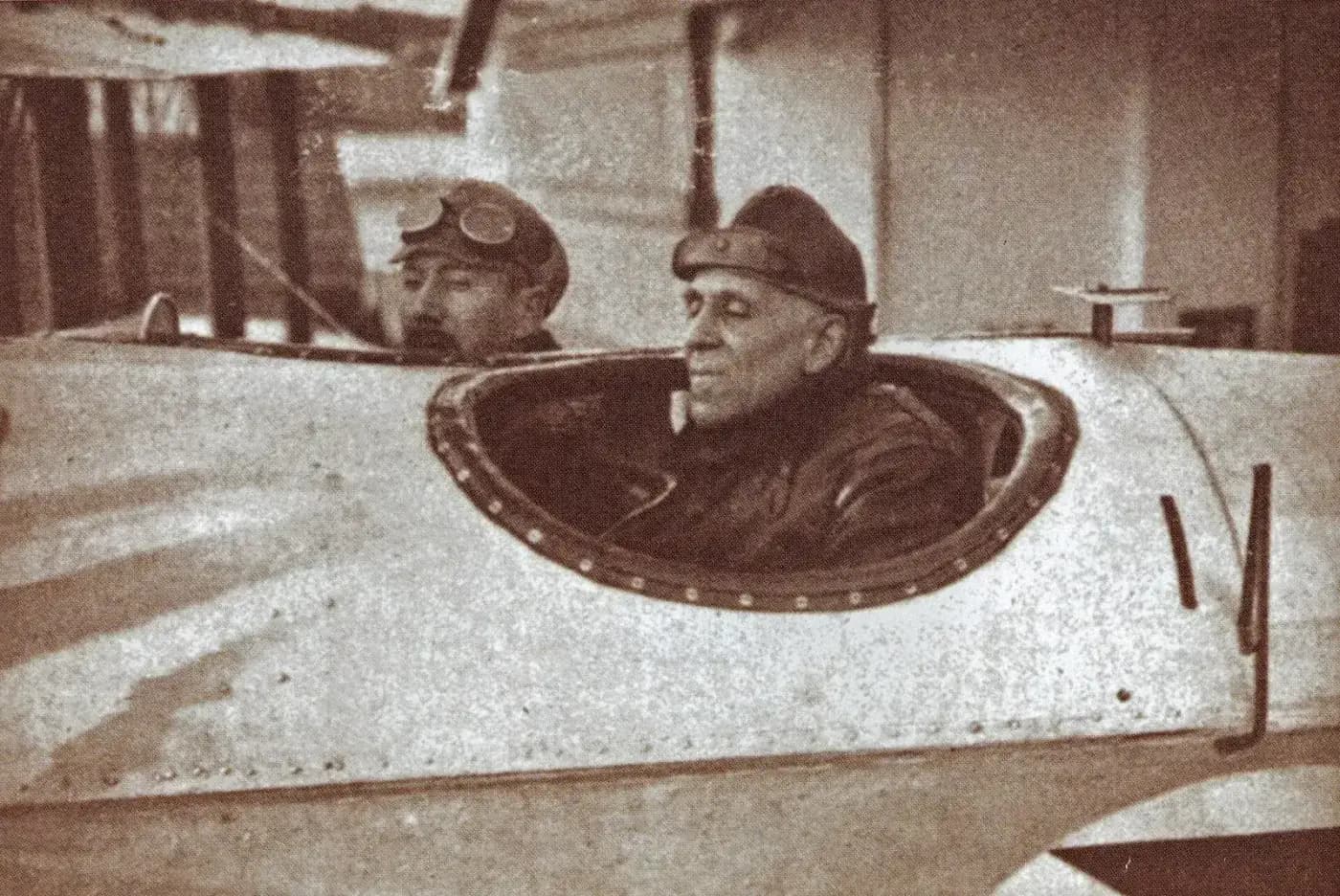Two airmen in leather helmets sit in the open cockpits of a biplane with a pale metal fuselage. The man in front is older, with grey hair and a calm expression; the man behind him has a moustache and flying goggles pushed up on his helmet.