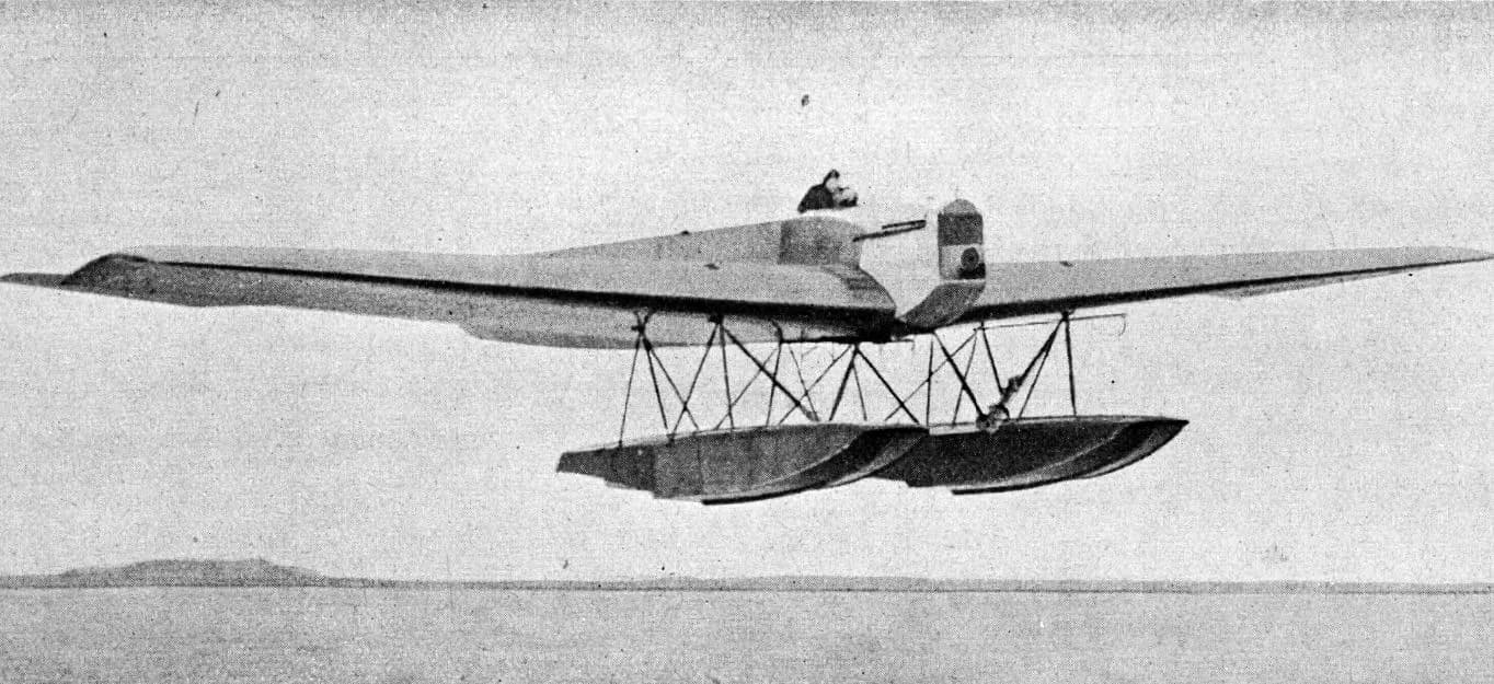 A long-winged single-engine monoplane with two large boxy floats flies low over flat water, with a pilot visible in the open cockpit and a low strip of land on the horizon.