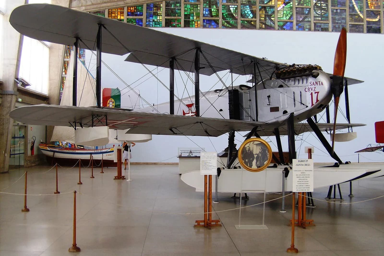A large silver biplane on white floats stands in a museum hall under a band of coloured glass windows. Its nose is painted “Santa Cruz 17” in red, a red cross is on the fuselage and a green and red Portuguese flag on the tail. A round framed photograph of two airmen stands in front of it.