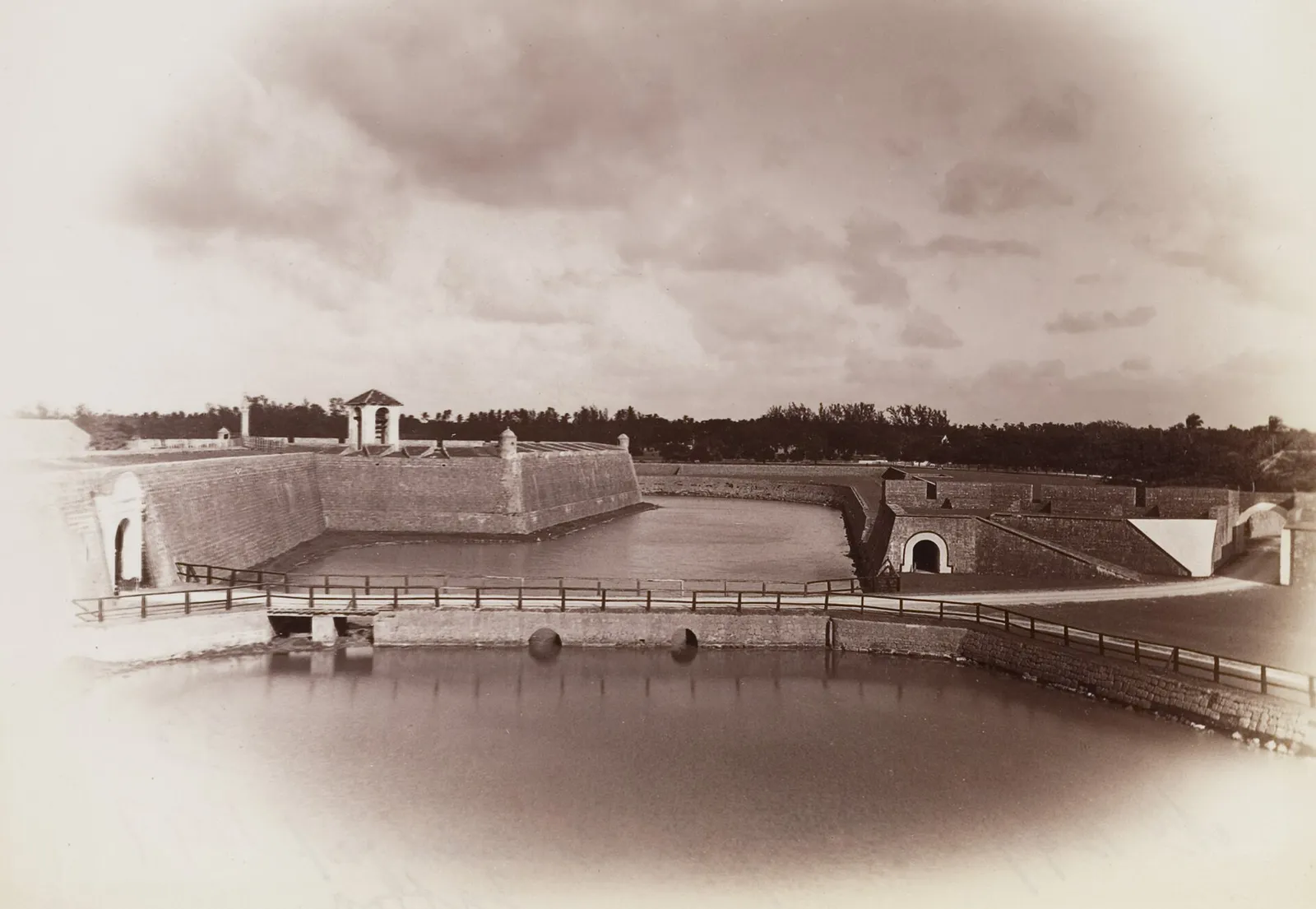 Sepia photograph of a wide moat of still water in front of low, sloping stone ramparts. A small roofed tower stands on the left bastion and arched gateways open in the walls, with a railed causeway and a pond in the foreground, under a cloudy sky.