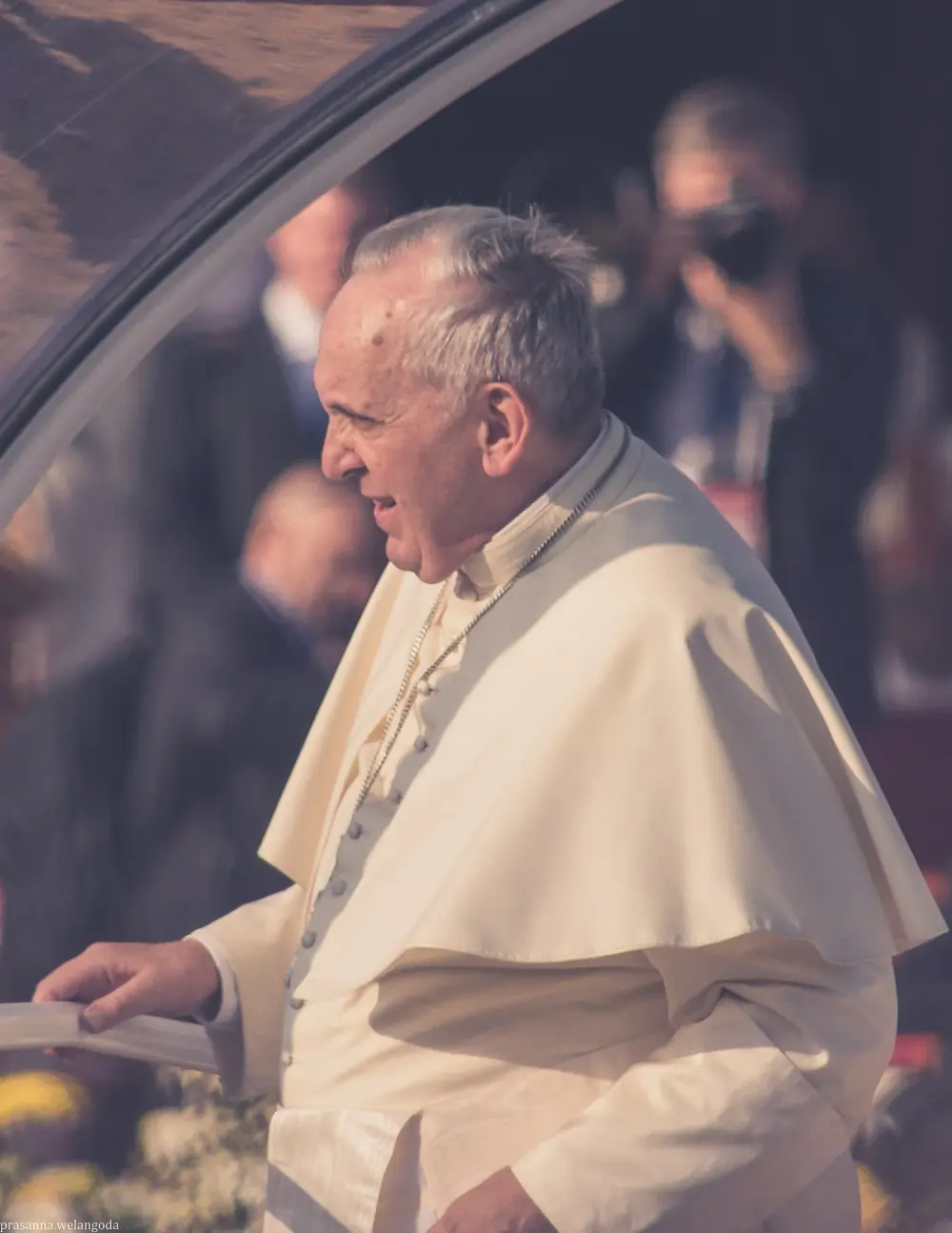 Pope Francis in a white cassock and short cape, smiling in profile beside the curved frame of his vehicle’s canopy, with a photographer and blurred onlookers behind him.