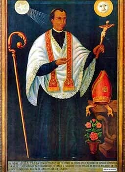 Painted portrait of a dark-haired man in a white lace surplice and gold-trimmed red stole, holding up a crucifix. A bishop’s crozier stands at the left of the picture and a mitre rests on a ledge at the right beside a pot of flowers, with a rayed star and a sun with a face in the upper corners and an inscription along the bottom.