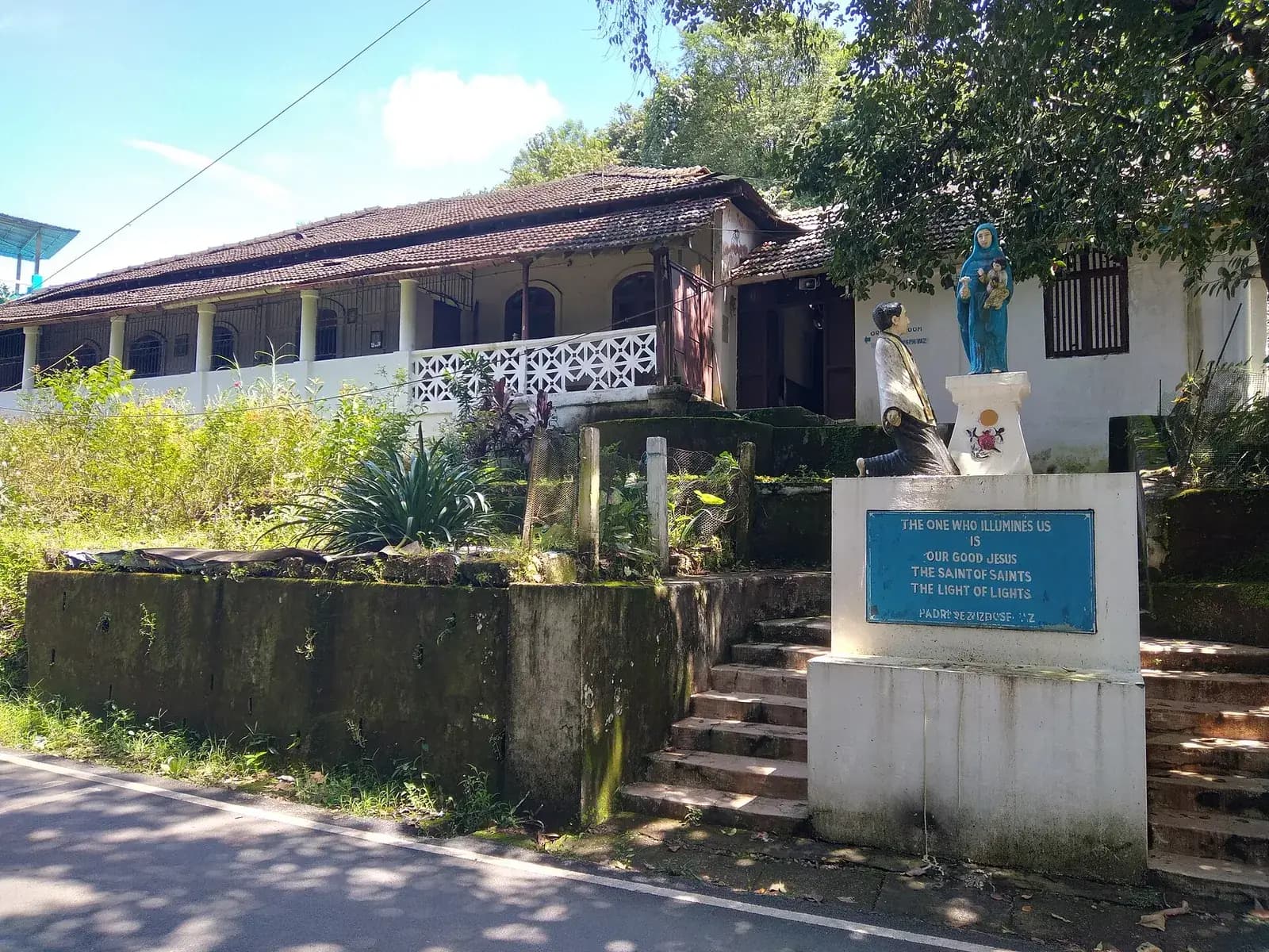 A long single-storey house with a tiled roof and a white balustraded veranda stands above a mossy retaining wall. In front, on a white pedestal, a statue of the Virgin Mary in blue rises behind a kneeling figure in white, above a blue plaque.