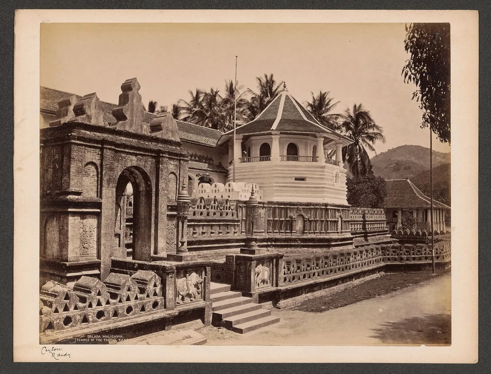 Albumen photograph of a carved stone gateway with an arched opening in the foreground, behind which rise a white octagonal pavilion with a pointed tiled roof and a low wall of carved stone balusters, with palm trees and hills behind.