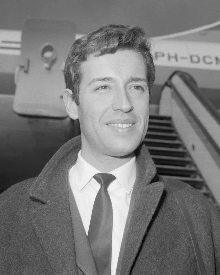 Black-and-white photograph of a smiling young man with wavy dark hair, in a white shirt, dark tie and heavy overcoat, standing in front of the boarding steps of an aircraft.