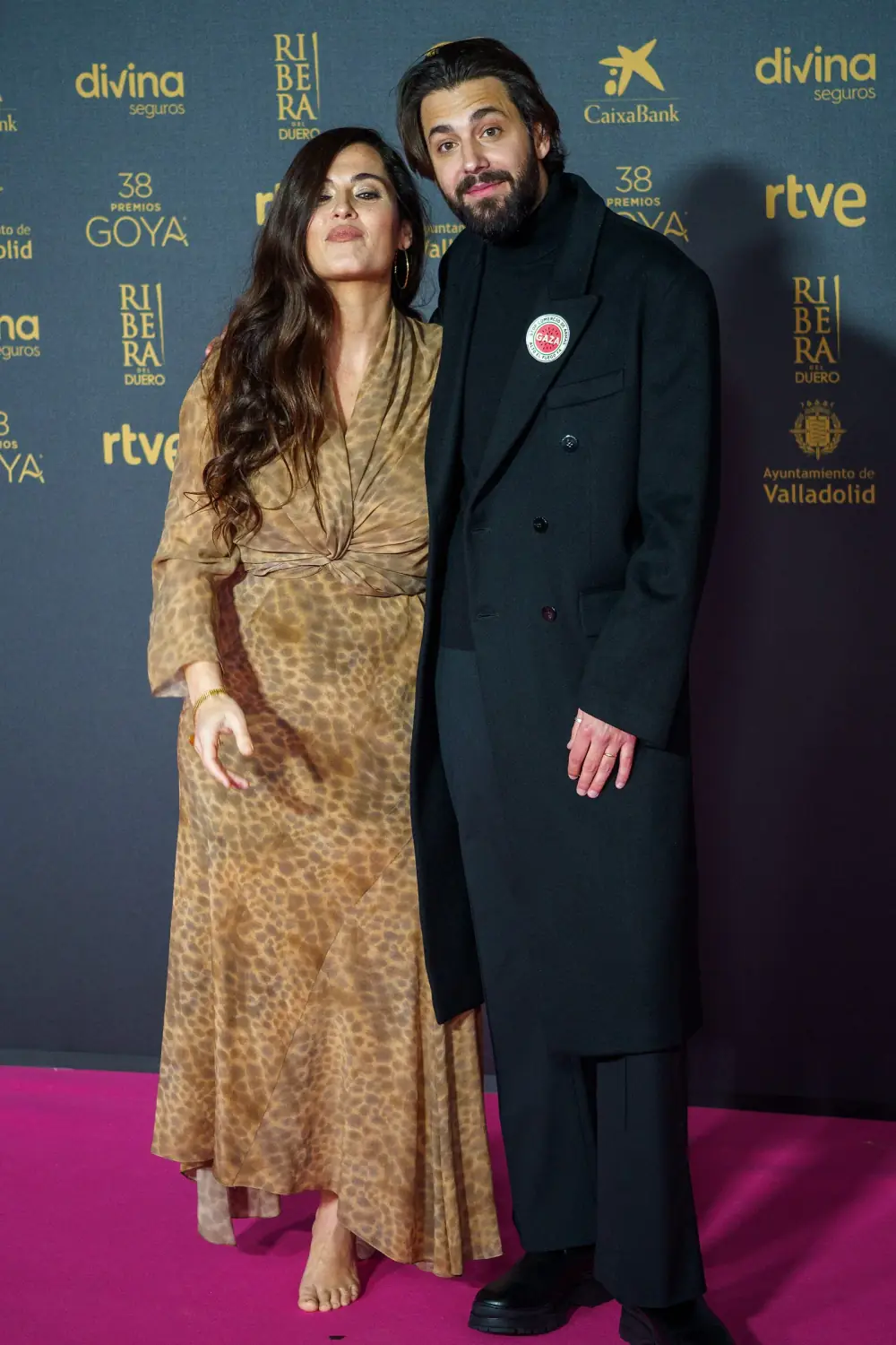 A woman with long dark wavy hair in a long leopard-print dress, barefoot, stands beside a bearded man in a long black overcoat with a round badge on the lapel, in front of a sponsor wall on a pink carpet.