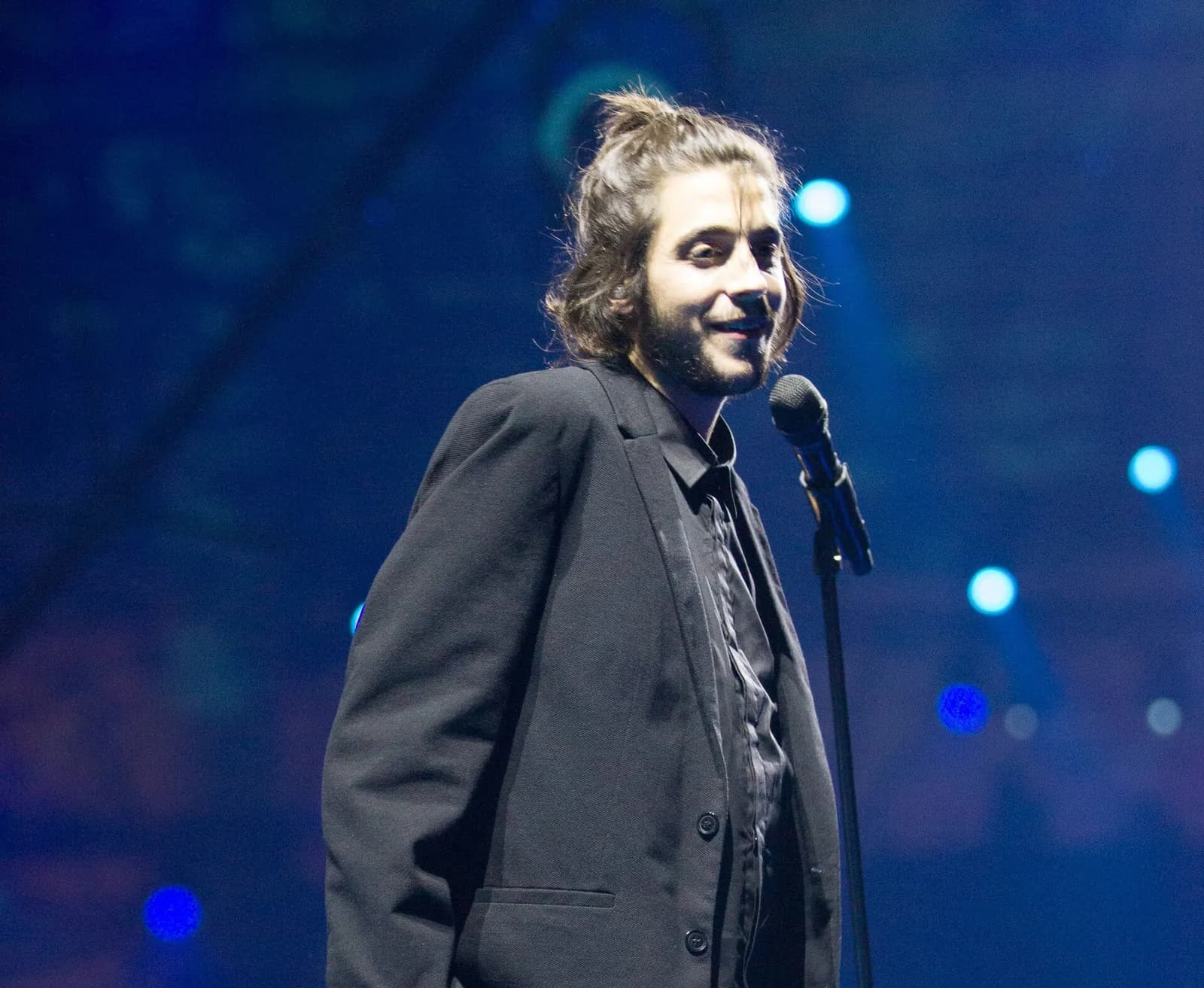 A young man with long hair tied up in a loose bun and a beard, wearing a loose black jacket over a black shirt, smiles beside a microphone stand on a dark stage lit with blue spotlights.