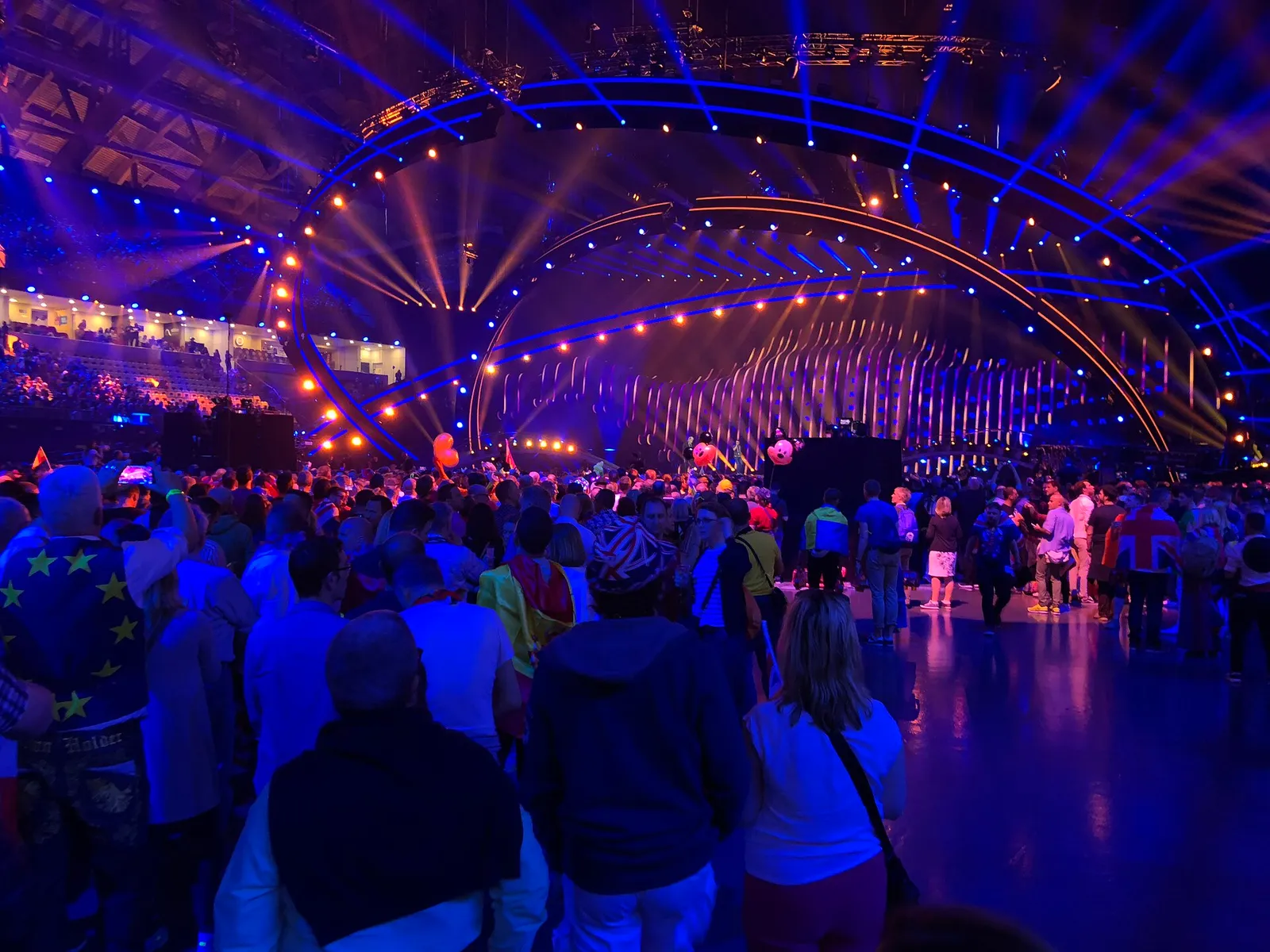 A crowd of fans, some wrapped in flags, fills the standing area of a dark arena in front of a large stage framed by curved rigs of blue and orange lights, with spotlights sweeping overhead.