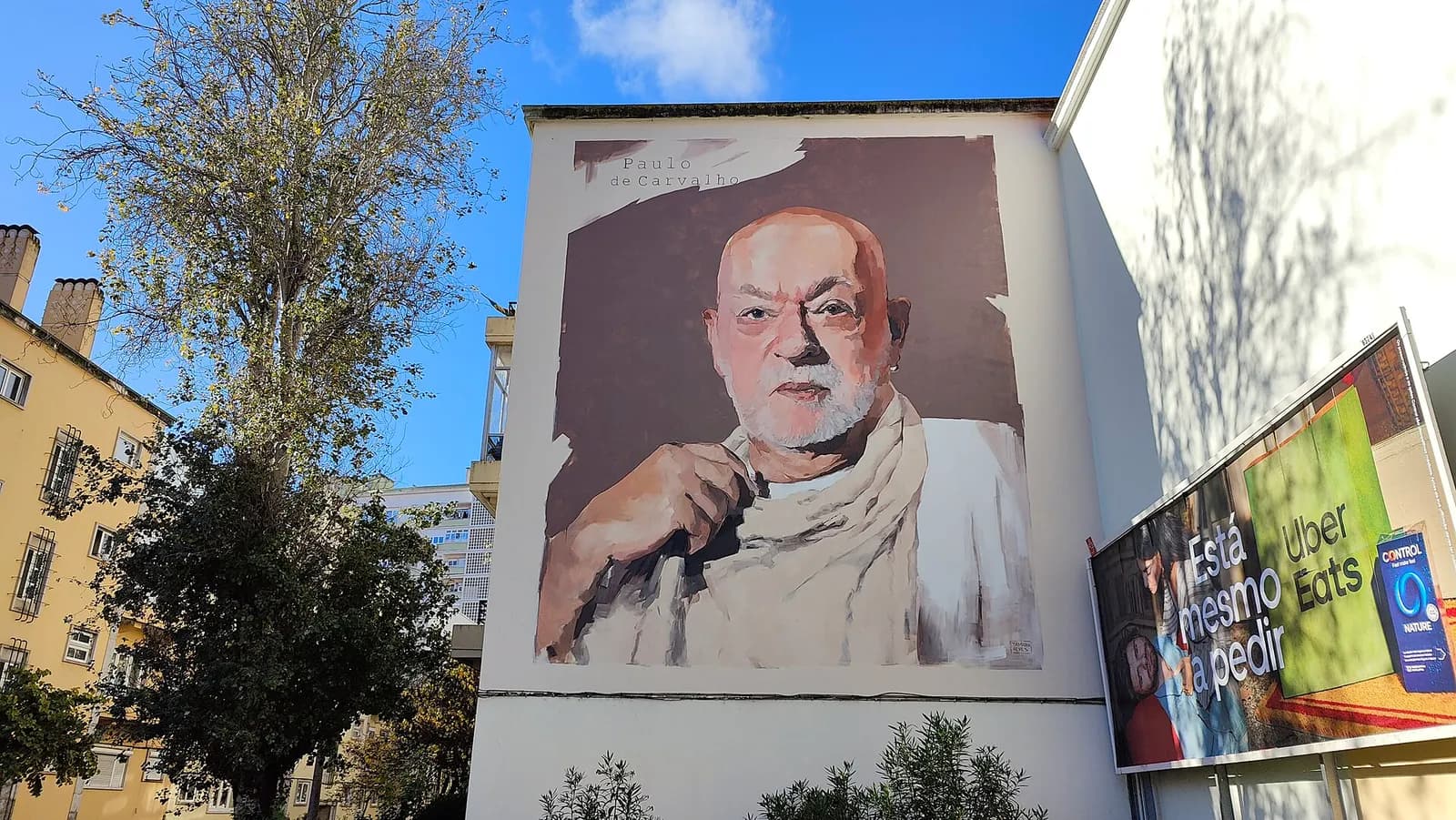 A large painted portrait on the end wall of a white building: a bald older man with a white beard, wrapped in a pale scarf, holding the scarf at his chest. The words Paulo de Carvalho are lettered in the top corner. Trees stand to the left and a billboard to the right.
