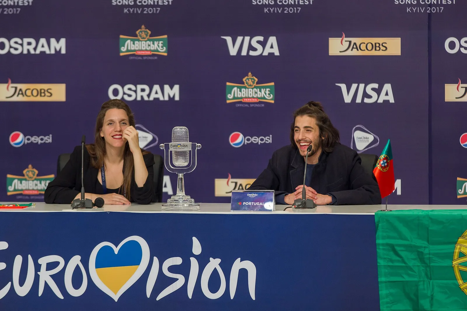 A smiling woman with long brown hair and a laughing man with his hair tied back sit at a table in front of a sponsor backdrop. Between them stands a glass trophy shaped like an old microphone; a Portuguese flag hangs from the table edge.