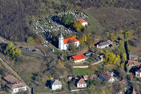 Aerial view of a hillside village in autumn: a white church with a small spire and a red roof, a cemetery of pale headstones on the slope behind it, and scattered houses with red and grey roofs among yellowing trees.