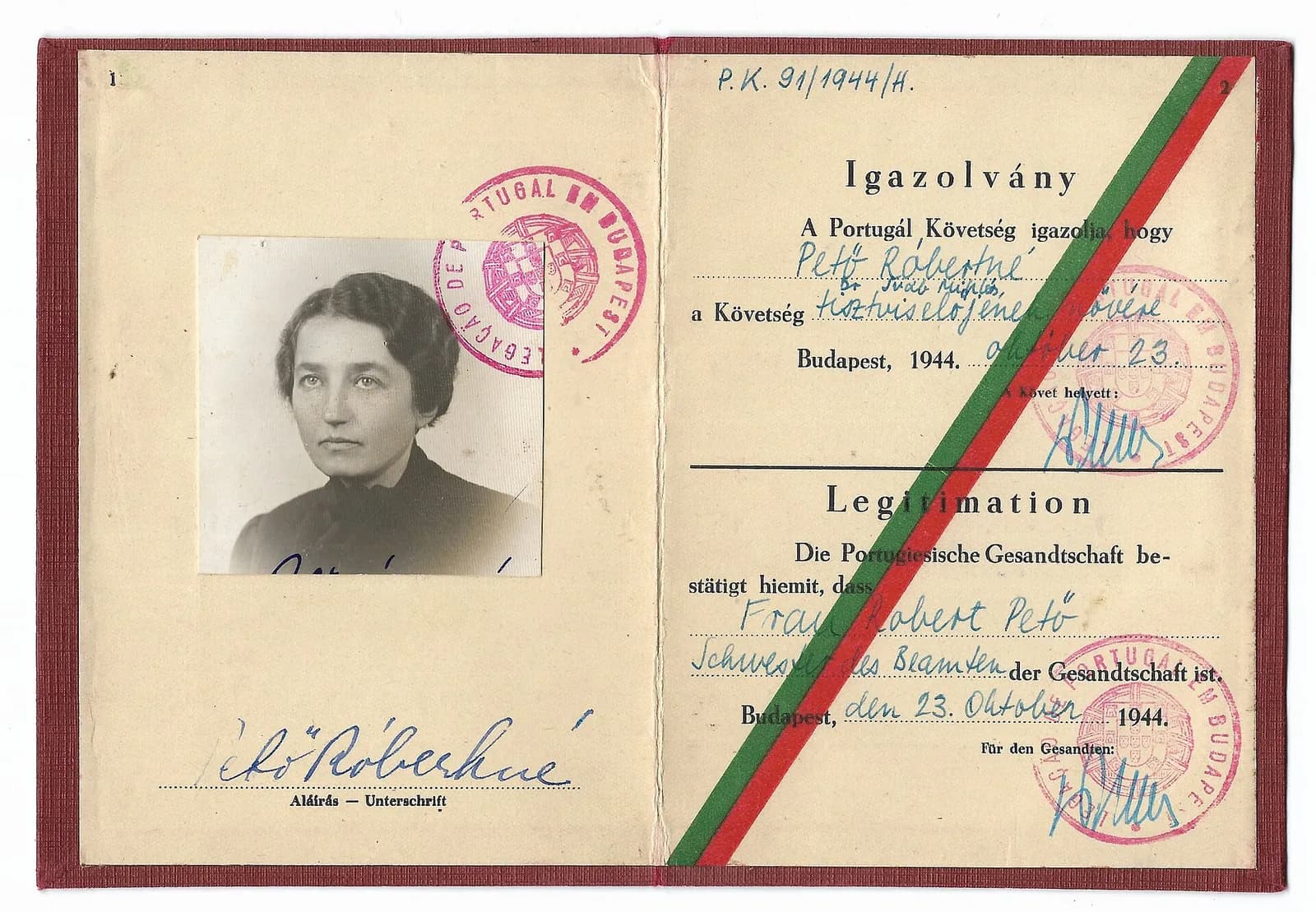 An open booklet with a red cloth cover. On the left page is a small black-and-white photograph of a dark-haired woman with a round red stamp of the Portuguese legation in Budapest and her signature below. The right page carries a diagonal green and red ribbon and a printed text in Hungarian and German, filled in by hand, dated Budapest, 23 October 1944, stamped and signed for the minister.