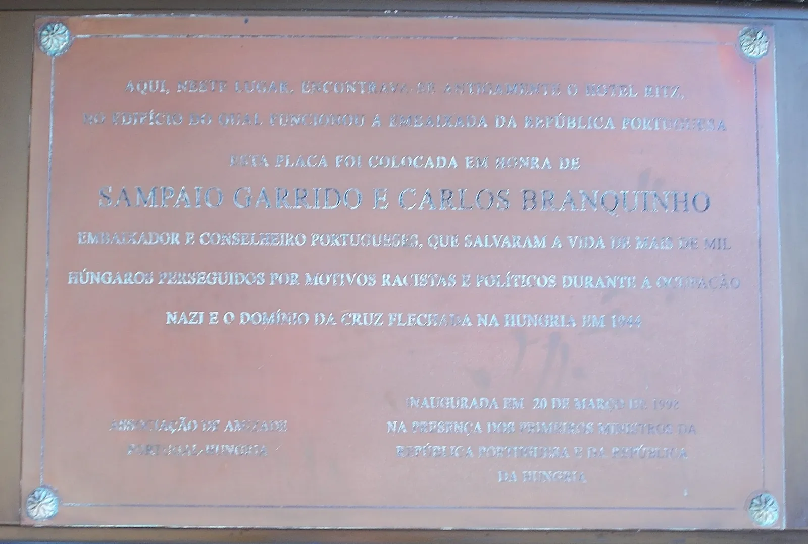 A rectangular copper-coloured plaque with raised capital letters in Portuguese and a small rosette in each corner. The largest line reads Sampaio Garrido e Carlos Branquinho.