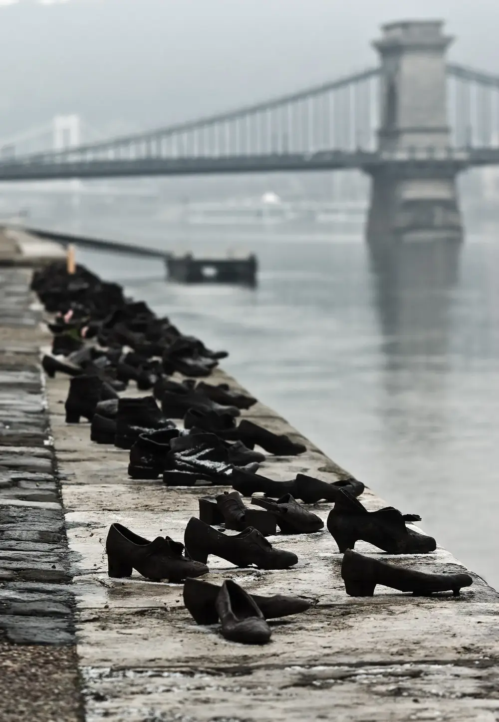 Rows of old-fashioned shoes cast in dark metal stand along the stone edge of a river embankment, men's boots and women's heeled shoes, with a suspension bridge fading into mist in the background.