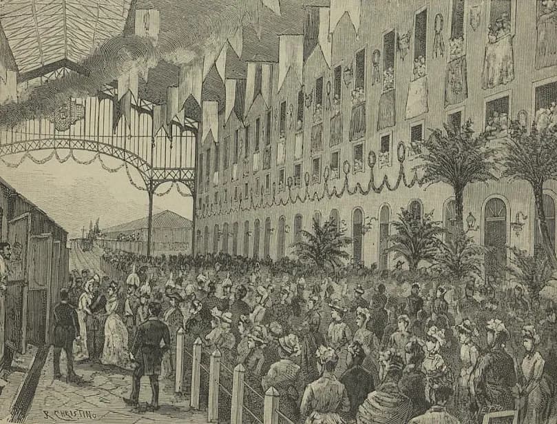 Engraving of a great crowd of men in top hats and women in bonnets packed along a platform under an arched iron roof. Flags and garlands hang along the wall of the station building and from its windows, where more people lean out, and potted palms line the platform. On the left a group steps down from a railway carriage.