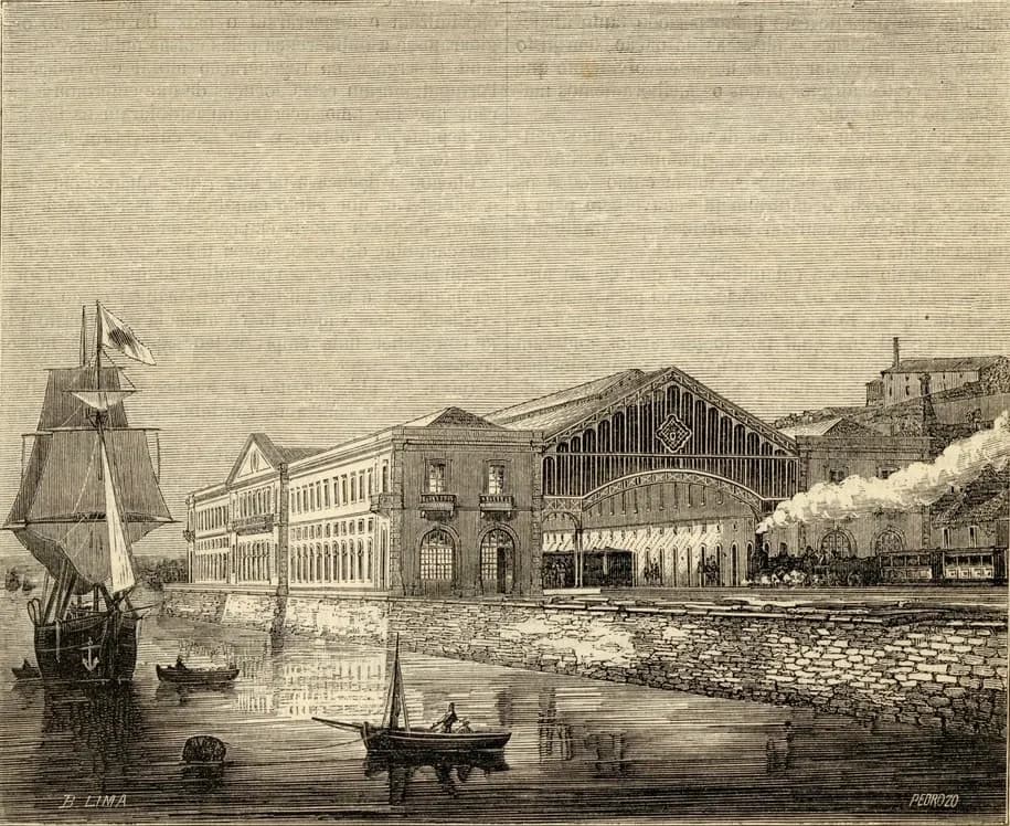 Engraving of a long two-storey building with a pedimented far end, standing on a stone quay at the water's edge. At its near end rises the tall glazed gable of an arched iron train shed, open to the tracks, where a steam train puffs smoke. A sailing ship and small boats float on the river in the foreground.