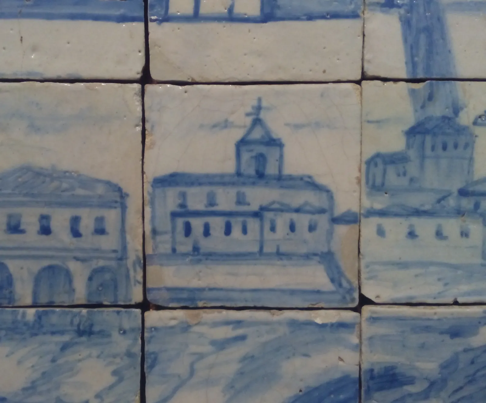 Detail of blue and white glazed tiles showing a plain two-storey building with a row of small windows and a bell tower topped by a cross, standing above the water's edge.