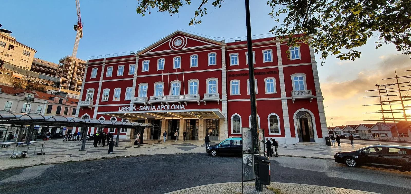 A three-storey building painted deep red with white trim, a pediment with a round clock at the top and the words Lisboa – Santa Apolónia above a glass canopy over the entrance. Cars and pedestrians are in the square in front, and the masts of a sailing ship rise on the right against an early-morning sky.