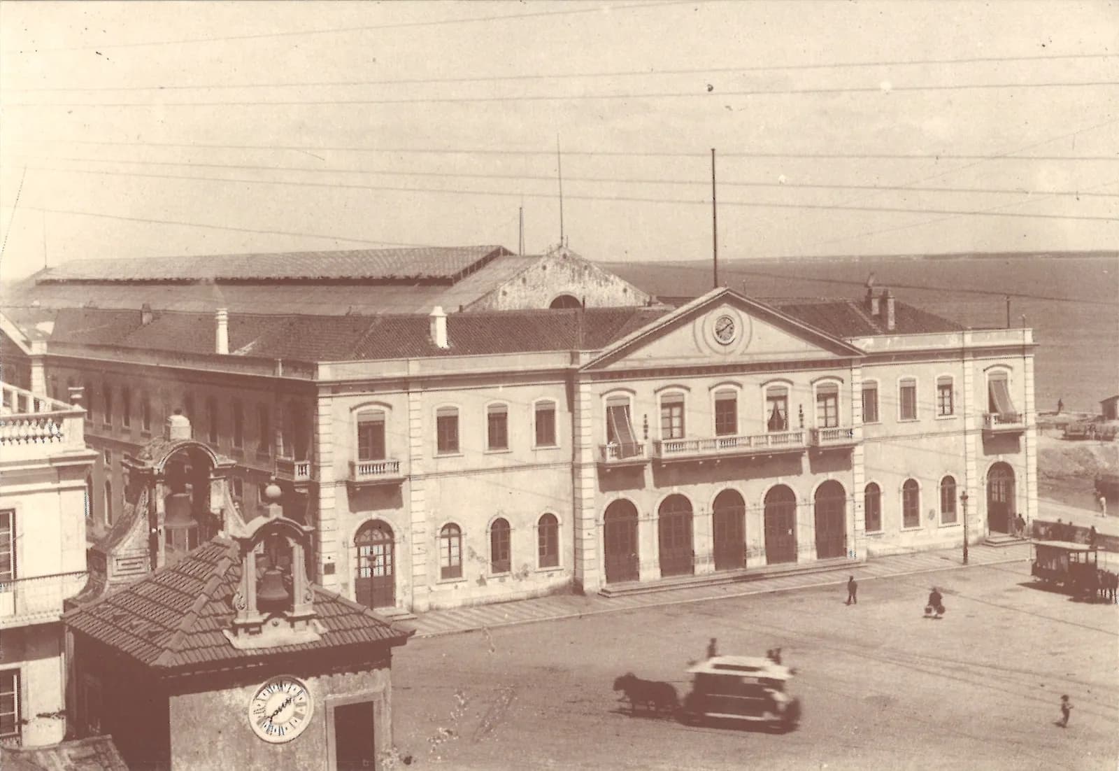 Old sepia photograph of a long two-storey station building with a central pediment holding a clock, five tall arched doors and balconies above. The long roof of a train shed rises behind it, with the river beyond. A horse-drawn vehicle crosses the empty square in front, and a small bell tower with a clock stands in the foreground.