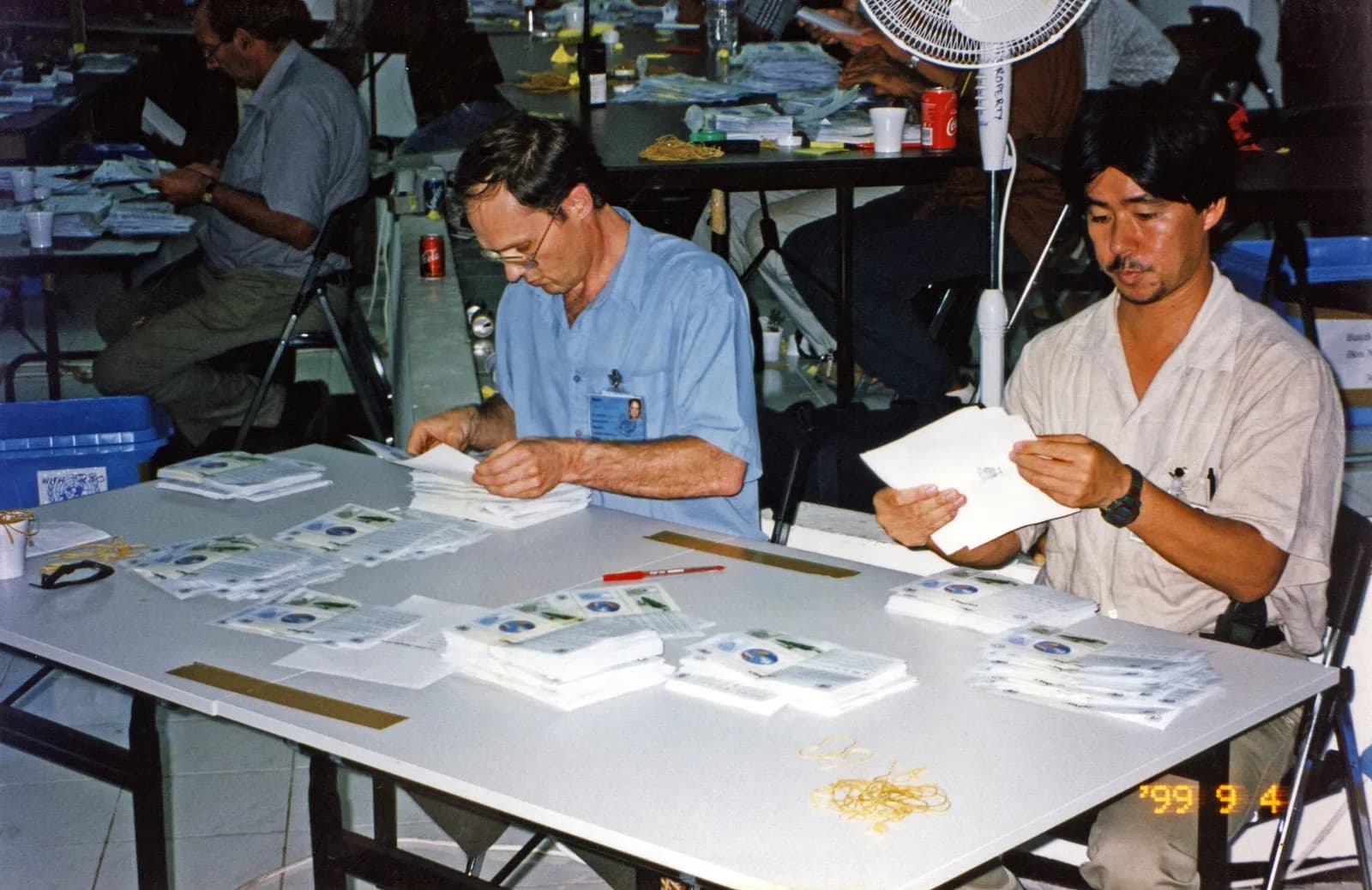 Two men sit at a white folding table sorting stacks of ballot papers printed with small coloured pictures, with other counters at tables behind them and a blue UN ballot box on the floor.