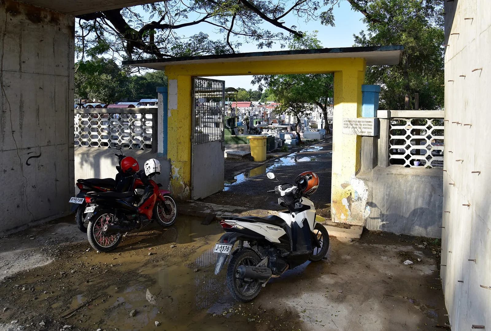 A yellow and blue concrete gateway with an open metal gate leads into a cemetery crowded with white tombs. Motorcycles are parked on the muddy ground in front of it.
