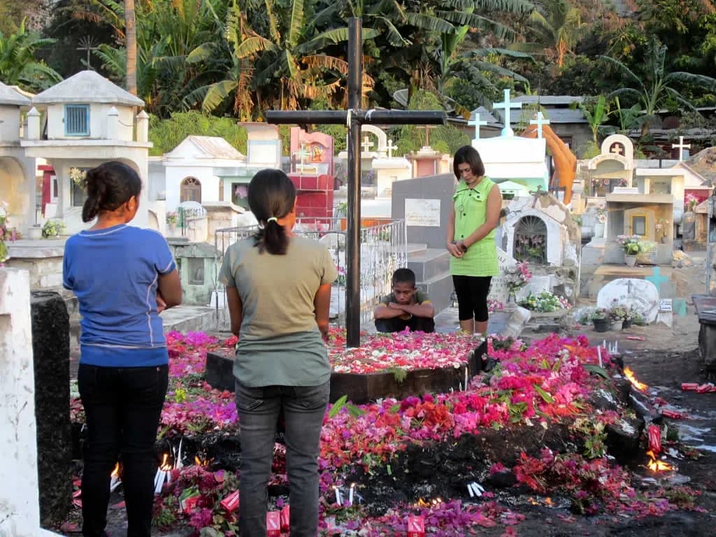 Three young people stand and a boy crouches beside a grave covered in red and pink petals and candles, under a tall dark cross, among white and pastel tombs.
