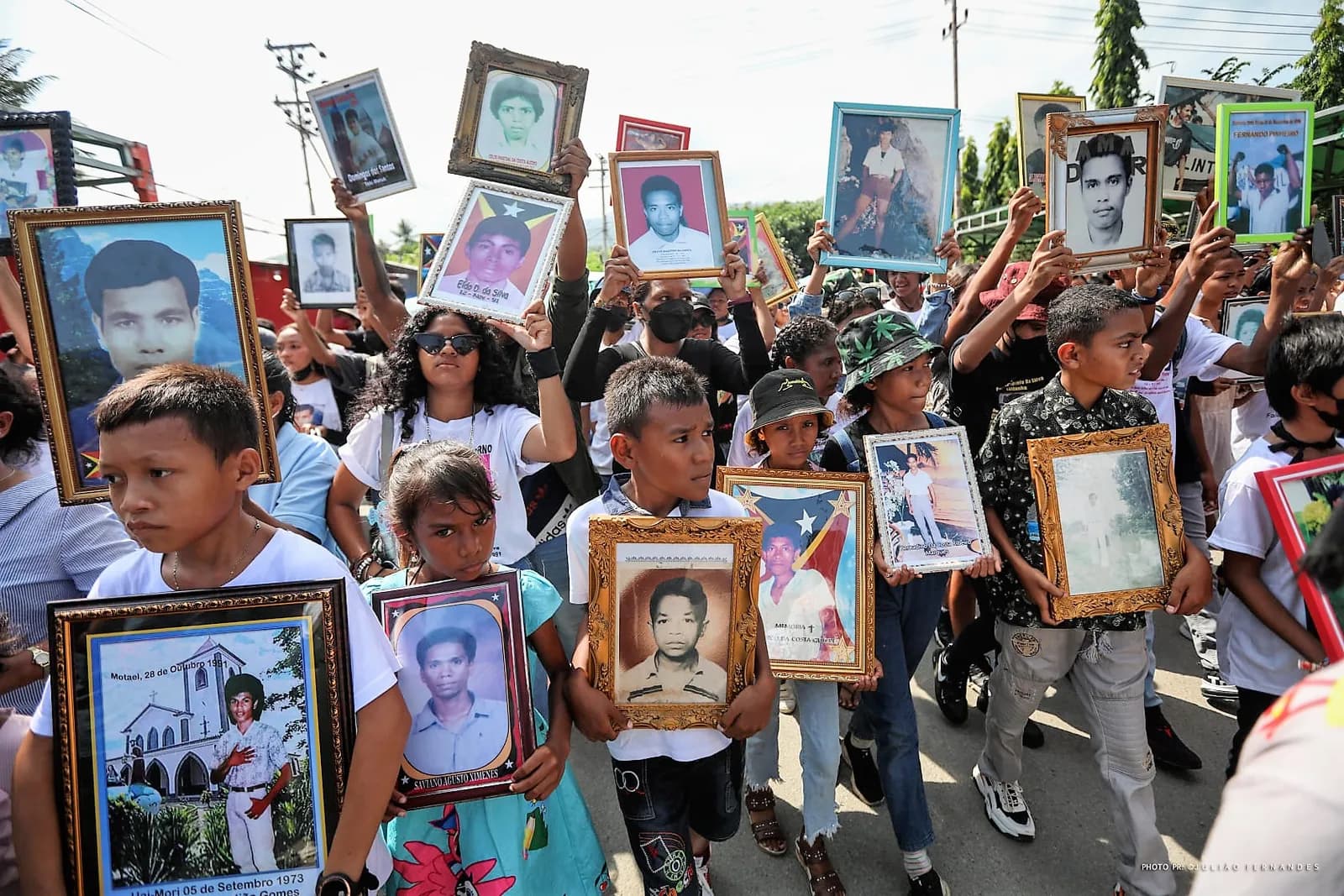 A crowd of children and young people walk down a street holding up framed photographs of young men. A boy in front carries a frame with a picture of a church and the words Motael, 28 de Outubro 1991.