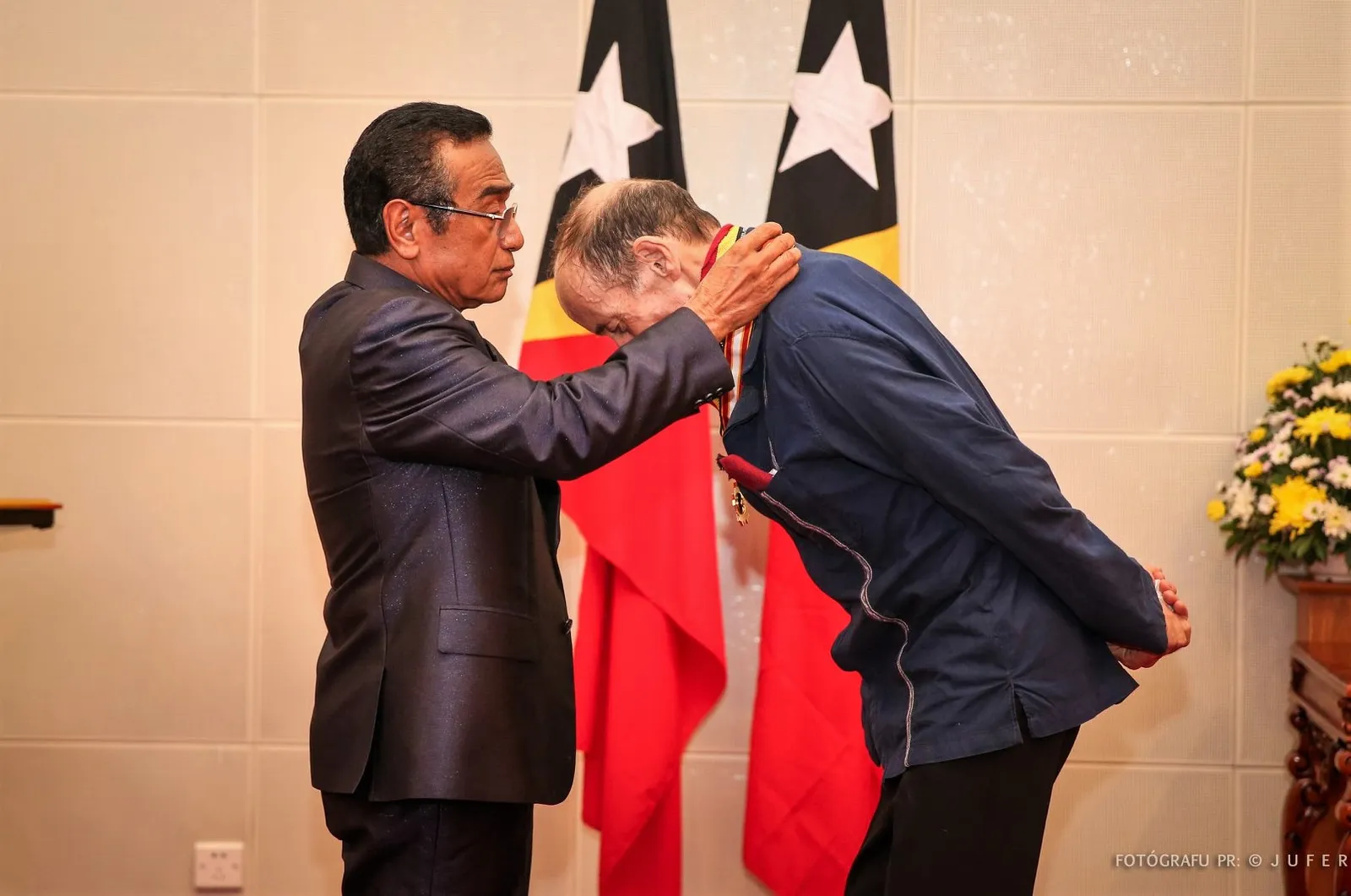 A thin, balding man in a navy jacket bows his head, hands behind his back, as a dark-haired man in glasses and a dark suit places a medal on a ribbon round his neck, in front of two Timor-Leste flags.