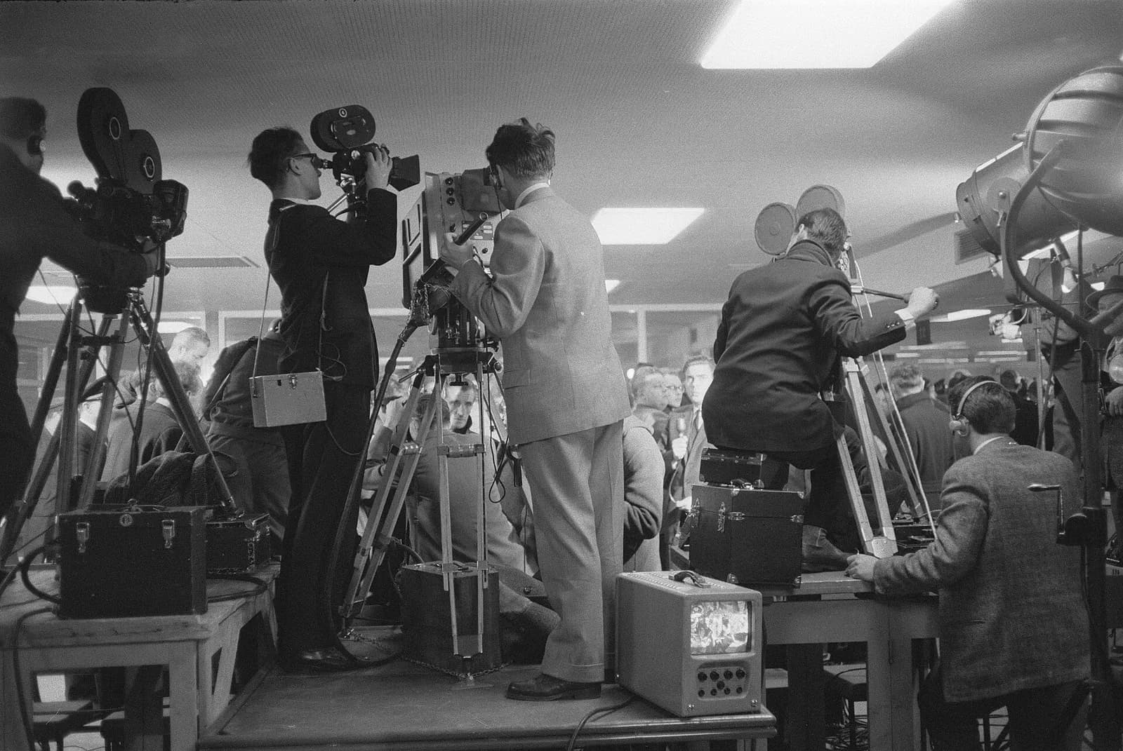 Black-and-white photograph of newsreel cameramen in suits standing on a raised platform in an airport hall, film cameras on tripods, a large lamp on the right and a small television monitor at their feet, with a crowd filling the hall beyond.