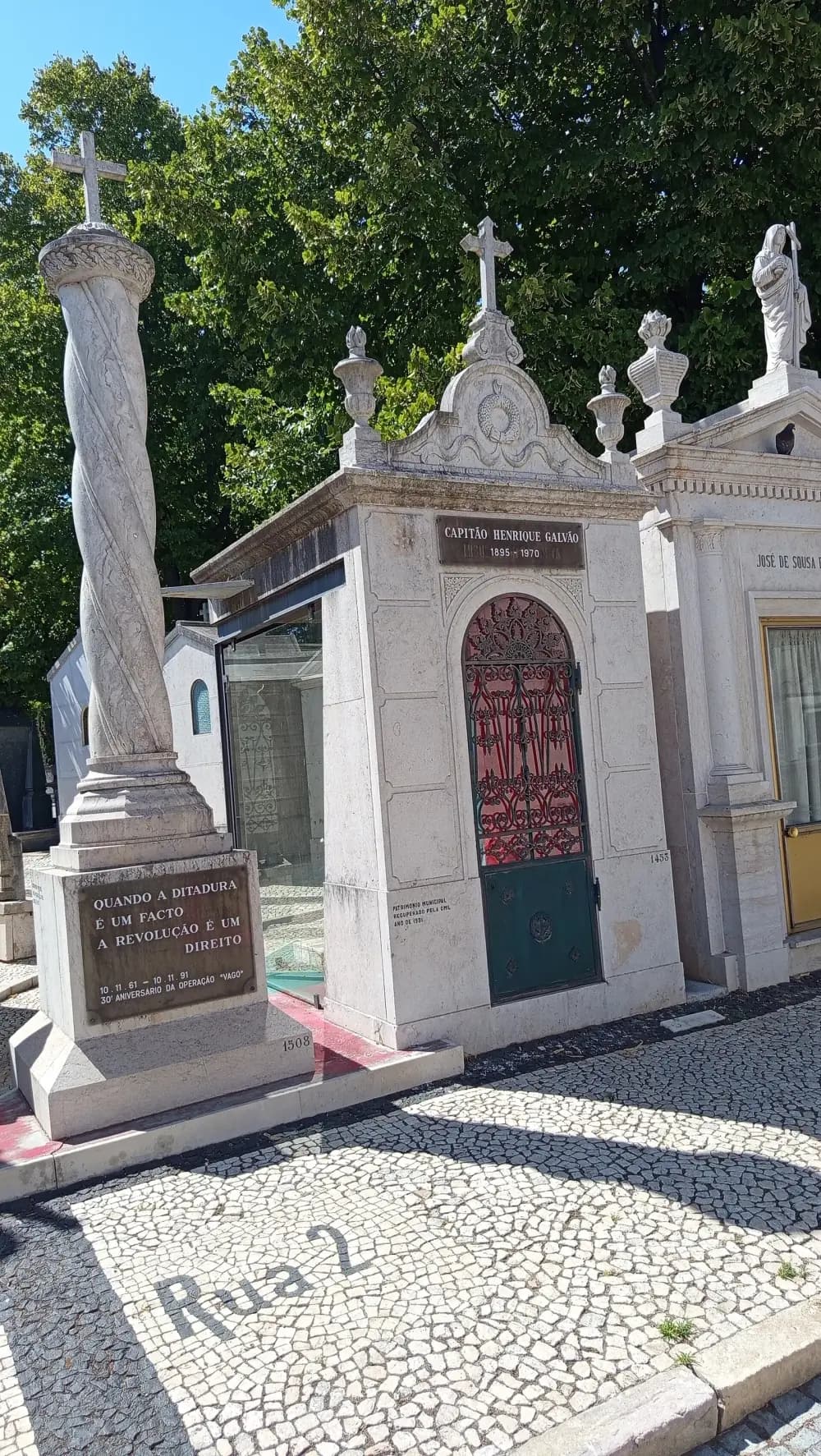 A small white stone mausoleum topped by a cross, with a bronze plaque reading Capitão Henrique Galvão 1895-1970 and a green and red iron door; beside it a twisted stone column with a plaque, on a cobbled cemetery street marked Rua 2.