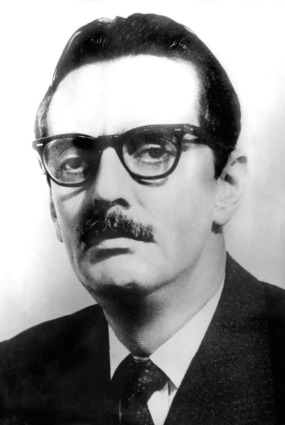 Black-and-white official portrait of a man with dark hair swept back, heavy black-framed glasses and a moustache, wearing a dark suit and patterned tie.