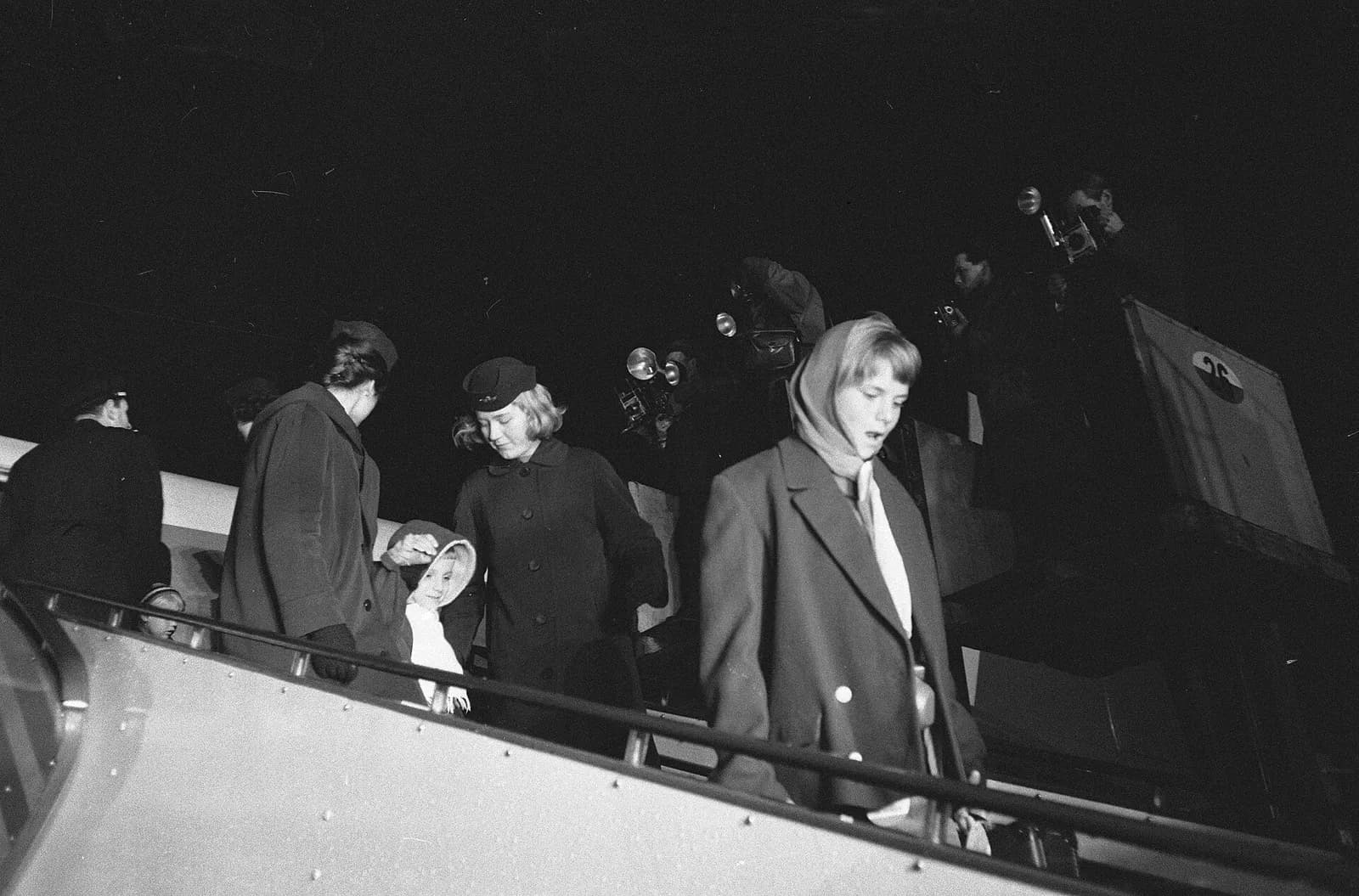 Black-and-white night photograph of passengers in winter coats walking down the steps of an aircraft: a girl in a headscarf in front, a young woman in a uniform cap beside a small child in a white-lined hood, and press photographers with flash cameras on a platform behind.